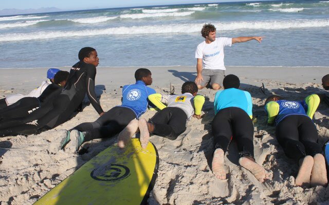 Take a first glance at this gaggle of teenagers stood round in a huddle on one of the most picturesque beaches in Cape Town and you’d be forgiven for thinking it was a bunch of mates about to catch some after-school surf. But get closer and you’d see there’s a coach stood among them, raising his voice so he can be heard over the crashing wave break, he says “so tell me, how does it feel to be pushed around?”